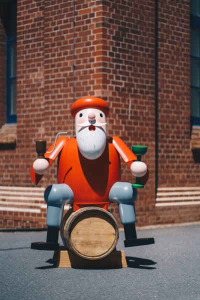 Ein roter lebensgroßer Räuchermann aus Holz sitzt auf einem Weinfass. Dieses steht vor einer Wand aus Backsteinen.